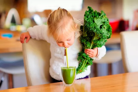 Una niña sujeta una col rizada en la mano mientras bebe un zumo de kale