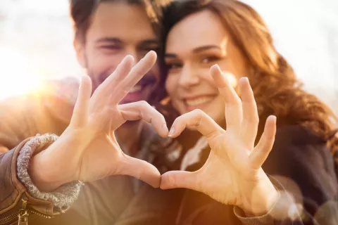 Pareja sonríe a la cámara formando con sus manos un corazón