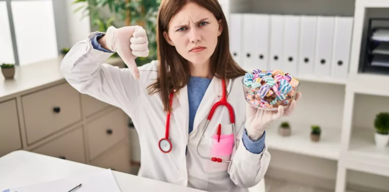 Joven doctora muestra caramelos y el pulgar hacia abajo Doctora sujeta un cuenco lleno de caramelos y hace un gesto de desacuerdo con el pulgar hacia abajo
