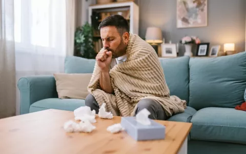 Temporada de gripe se adelanta Hombre de mediana edad en su sofá arropado con una manta, con tos y pañuelos en la mesa