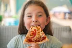 Dar ultraprocesados a los niños pone en riesgo su salud cardiometabólica Una niña joven comiendo una porción de de pizza