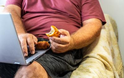 Un adulto sentado en el sofá trabajando con el portátil y comiendo comida no saludable